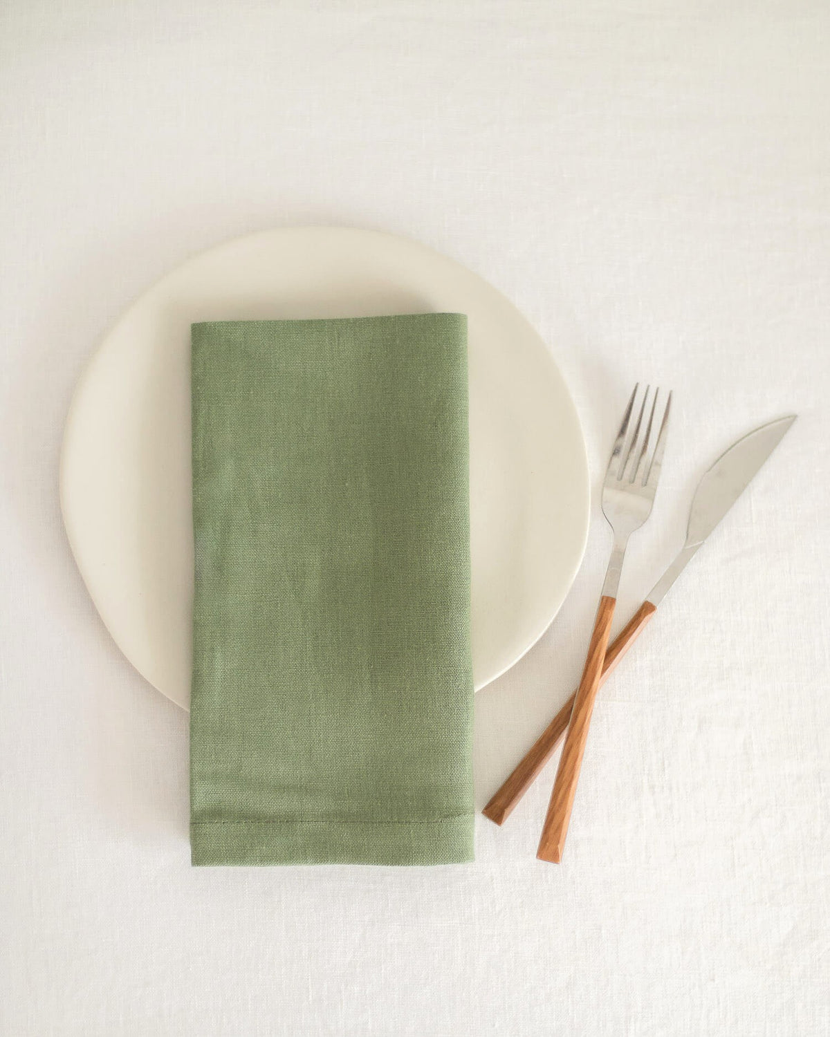 ava and ava ph sage green pure linen table napkin on off-white plate, folded into rectangular shape, with wooden spoon and fork