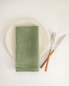 ava and ava ph sage green pure linen table napkin on off-white plate, folded into rectangular shape, with wooden spoon and fork