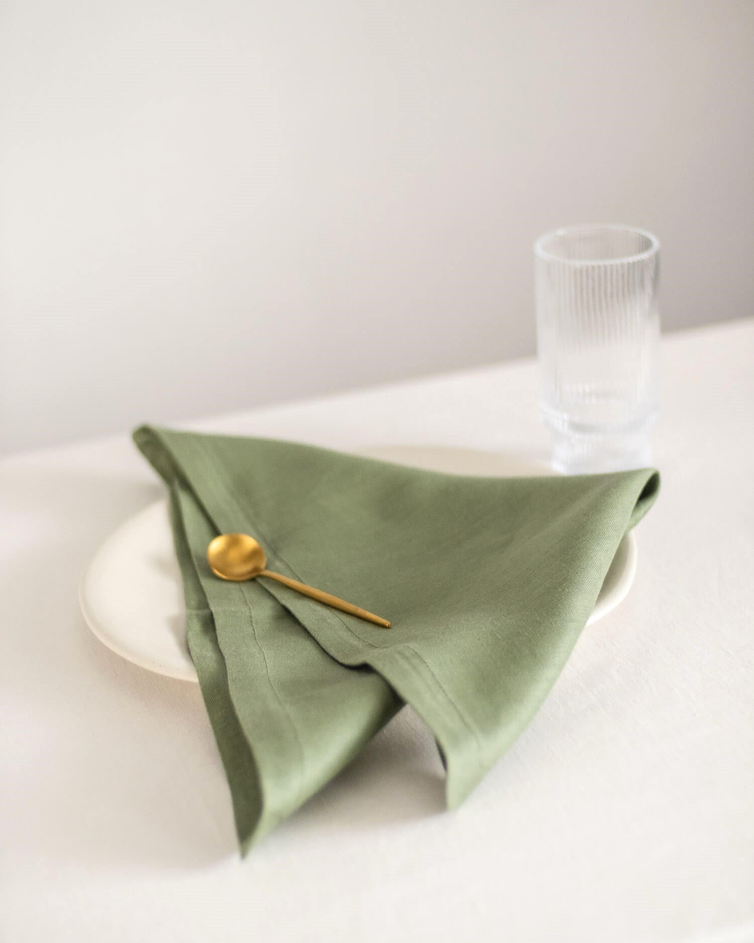 ava and ava ph sage green pure linen table napkin on off-white plate, folded into triangular shape, with wooden golden spoon and water glass
